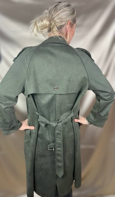 Model set bagfra iført olivengrøn trenchcoat i blødt ruskindslook med klassisk rygskæring, skulderflap og markeret taljebælte – et elegant silhuetspil mellem struktur og bevægelse, der balancerer det rå og det raffinerede.