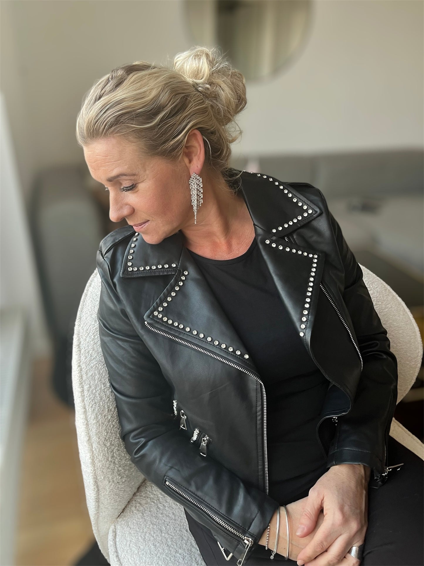 Model siddende afslappet på en stol iført sort faux læder bikerjakke med markeret bælte i taljen, opsmøgede ærmer og let åbent snit – et casual, men powerfuldt look, hvor rå attitude får et feminint twist.