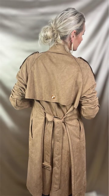 Model set bagfra iført olivengrøn trenchcoat i blødt ruskindslook med klassisk rygskæring, skulderflap og markeret taljebælte – et elegant silhuetspil mellem struktur og bevægelse, der balancerer det rå og det raffinerede.